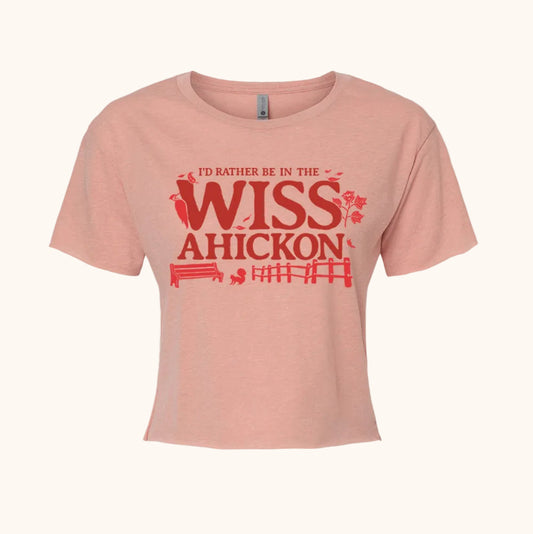 Pink crop top with red and maroon text 'I'd Rather Be in the Wissahickon' surrounded by a woodpecker, flowers, a bench, and fence on a beige background. 