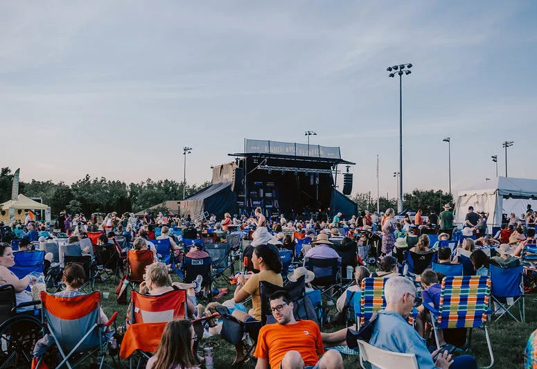 Outdoor concert with people sitting in chairs and on blankets facing a stage.