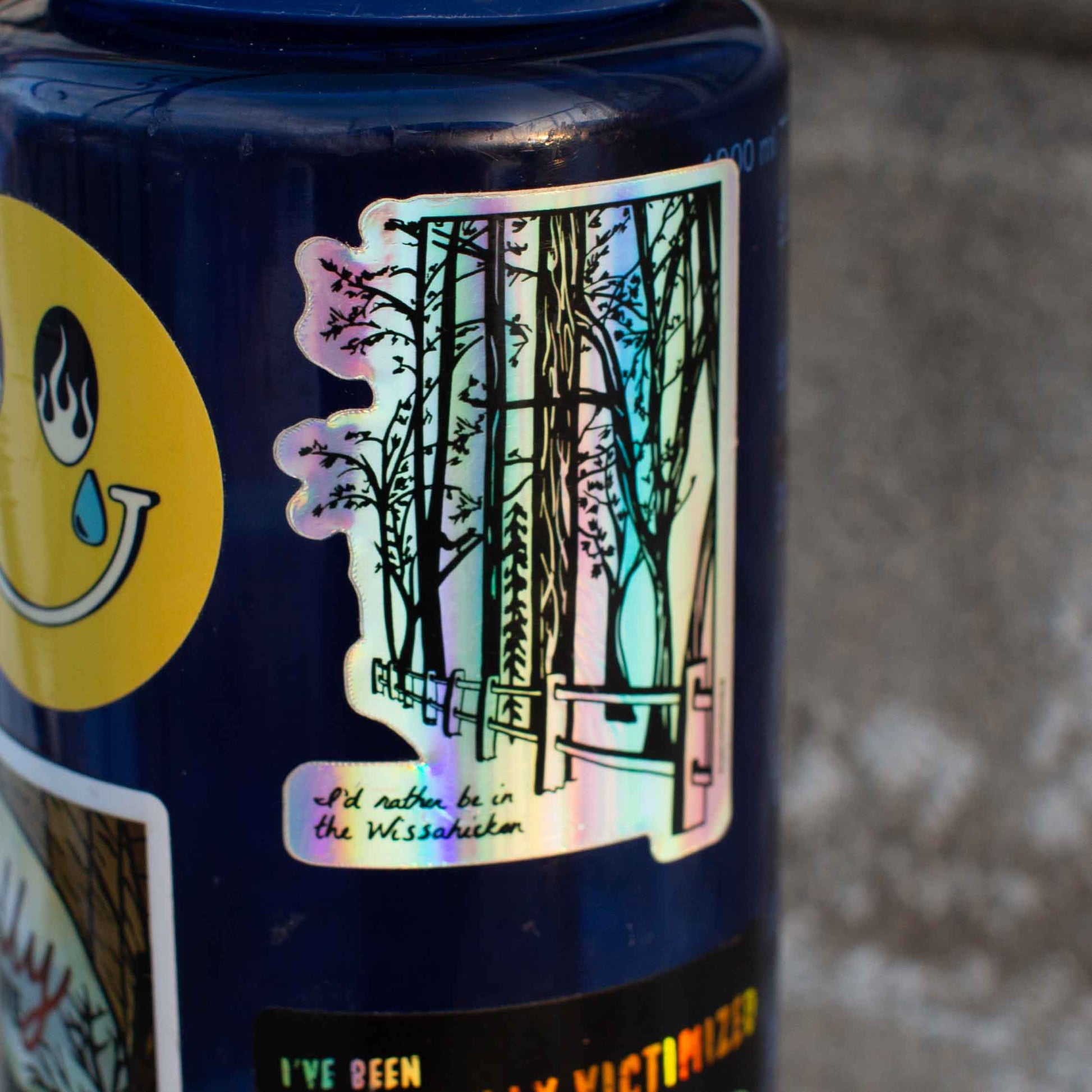 A holographic sticker with an illustration of the Wissahickon Valley Park, featuring trees and a pathway, on a blue water bottle
