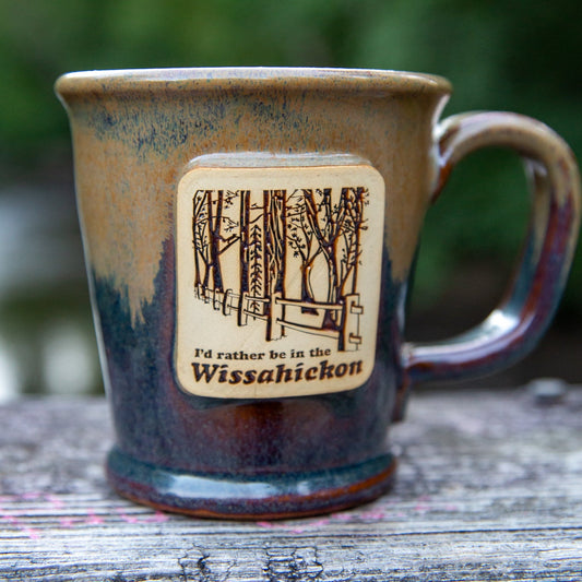 Brown gradident ceramic mug with text 'I'd Rather be in the Wissahickon' with fence and tree line illustration on wooden table.