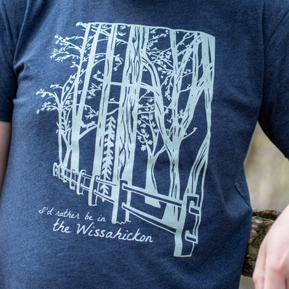 Young man wearing a dark blue t-shirt with a light green fence and forest with text 'I'd rther be in the Wissahickon' while standing outdoors.