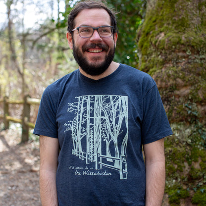 Young man wearing a dark blue t-shirt with a light green fence and forest with text 'I'd rther be in the Wissahickon' while standing outdoors and smiling.