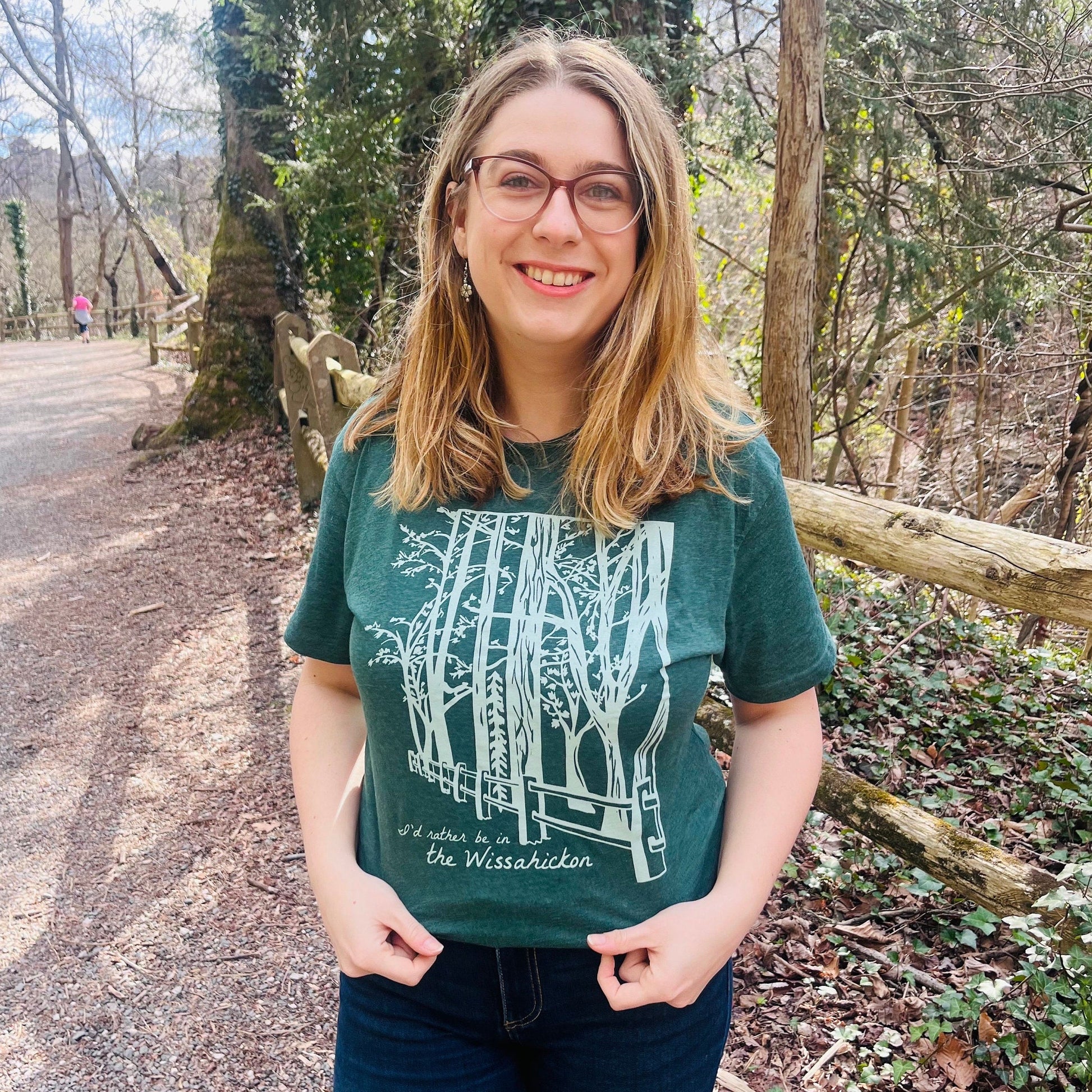 Young woman wearing a green t-shirt with a light green fence and forest with text 'I'd rther be in the Wissahickon' while standing outdoors and smiling.
