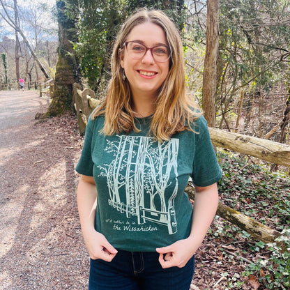 Young woman wearing a green t-shirt with a light green fence and forest with text 'I'd rther be in the Wissahickon' while standing outdoors and smiling.