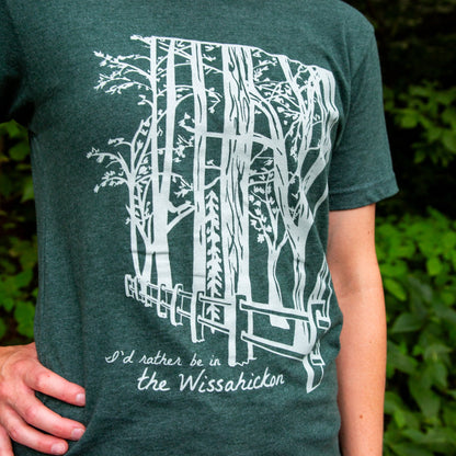 Young woman wearing a green t-shirt with a light green fence and forest with text 'I'd rther be in the Wissahickon' while standing outdoors.