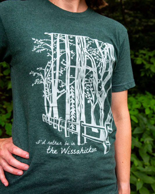 Young woman wearing a green t-shirt with a light green fence and forest with text 'I'd rther be in the Wissahickon' while standing outdoors.