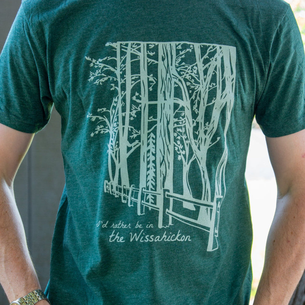 Young man wearing a green t-shirt with a light green fence and forest with text 'I'd rther be in the Wissahickon' while standing outdoors.