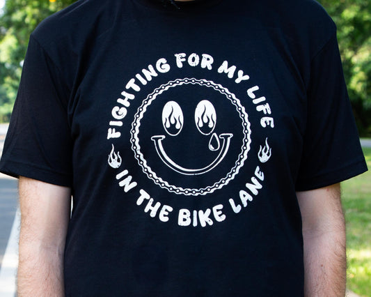 Black Tshirt with a White Illustration of a happy face with Flames in the Eyes that says "Fighting for my life in the bike lane " around the illustration 