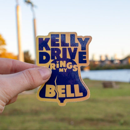 A blue and yellow waterproof vinyl sticker with the text 'KELLY DRIVE RINGS MY BELL' designed in the shape of a bell.