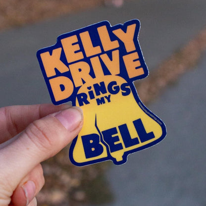 Hand holding a Librity bell shaped orange and blue sticker with 'Kelly Drive Rings against a blurred Kelly Drive background.
