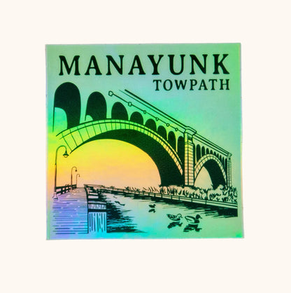 A sticker with a holographic design featuring the Manayunk Towpath in Philadelphia, with a bridge, duck, and fish illustrated on it.